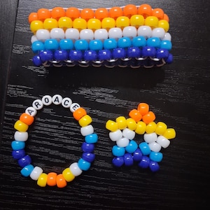 May include: A set of three handmade pony bead accessories including a wide rectangular cuff, a circular bracelet with the white letter beads spelling AROACE, and a star-shaped charm, all featuring horizontal stripes of orange, yellow, white, light blue, and dark blue.