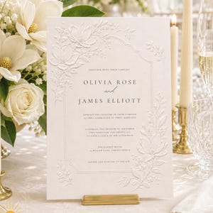 May include: A white wedding invitation card featuring embossed floral details and black printed text reading Olivia Rose and James Elliott, displayed upright on a gold stand. The card is surrounded by white flowers, candles, and glassware.