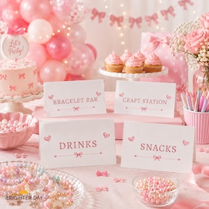 Op de afbeelding: Vier witte rechthoekige bordjes met roze tekst en strikillustraties, gelabeld Bracelet Bar, Craft Station, Drinks en Snacks. De bordjes staan op een tafel met een roze taart, cupcakes, kralen en roze ballonnen op de achtergrond.