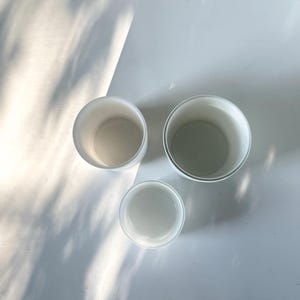 May include: Three white ceramic containers of varying sizes, stacked on top of each other. The containers are round and have a smooth, glossy finish.