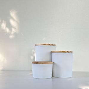 May include: Three white ceramic candle jars with wooden lids. The jars are stacked on top of each other, with the largest jar on the bottom and the smallest jar on top.