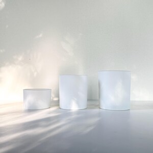 May include: Three white cylindrical candle jars of varying sizes. The jars are made of frosted glass and have a smooth, matte finish.