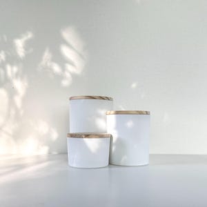 May include: Three white ceramic candle jars with wooden lids. The jars are stacked on top of each other, with the largest jar on the bottom and the smallest jar on top.