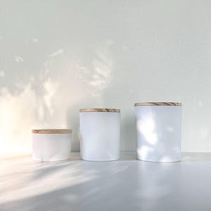 May include: Three white ceramic jars with wooden lids. The jars are different sizes, with the smallest jar in the front and the largest jar in the back. The jars are all white and have a smooth finish.