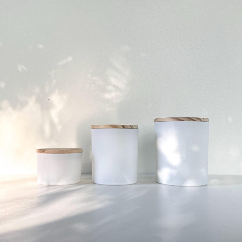 May include: Three white ceramic jars with wooden lids. The jars are different sizes, with the smallest jar in the front and the largest jar in the back. The jars are all white and have a smooth finish.