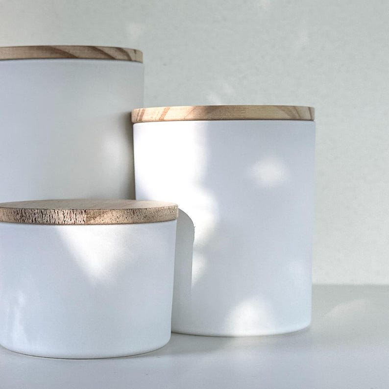 May include: Three white ceramic candle jars with wooden lids. The jars are different sizes and are arranged in a row from largest to smallest.