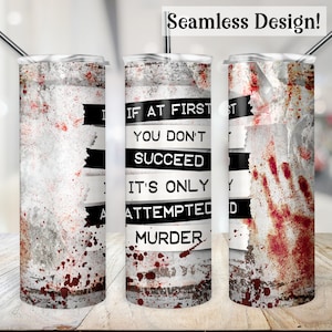 May include: A white tumbler with a black and white graphic that says "If at first you don't succeed it's only attempted murder". The tumbler has a red splatter pattern and a red handprint.