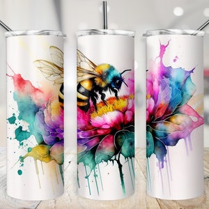Puede incluir: Una ilustración de estilo acuarela de una abeja en una flor con salpicaduras de pintura de colores. La abeja es amarilla y negra con detalles azules. La flor es rosa, morada y azul con hojas verdes. La imagen está impresa en un vaso blanco.