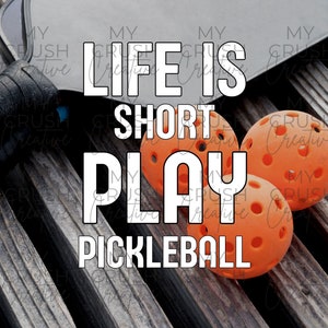K&ouml;nnte beinhalten: Ein schwarzer Pickleball-Schl&auml;ger mit dem Text "Life is Short Play Pickleball" in wei&szlig;en Buchstaben. Drei orangefarbene Pickleball-B&auml;lle befinden sich im Vordergrund.