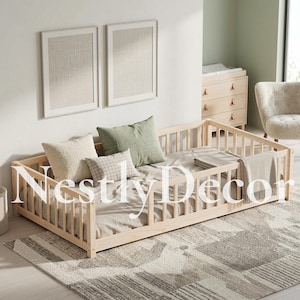 May include: A light-colored wooden floor bed frame featuring a slatted railing on three sides. The bed is dressed with a beige mattress, a textured cream pillow, a sage green pillow, a small plaid pillow, and a light gray throw blanket.