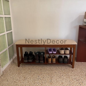 May include: A wooden shoe bench with a beige upholstered seat and two lower shelves for storage. The bench features a warm brown finish and is labeled with the text NestlyDecor. Multiple pairs of shoes are organized on the shelves below.