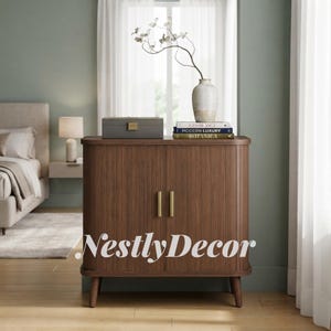 May include: A dark wood cabinet with two fluted doors, vertical slats, and two vertical brass handles. It stands on four tapered legs. On top sits a gray box, a white vase with branches, and three stacked books labeled Casablanca, Modern Luxury, and Botanica.