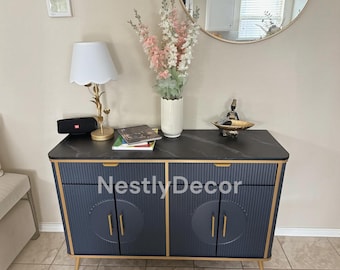 May include: A navy blue sideboard with a black marble-patterned top, gold trim, and vertical fluted cabinet doors featuring circular accents. The piece has gold handles and stands on four tapered legs. The text NestlyDecor is visible on the front.