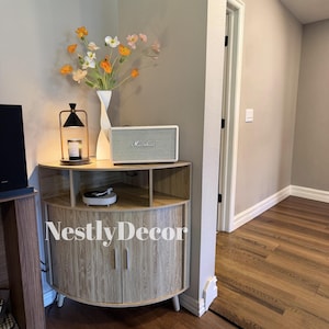 May include: A light wood corner cabinet with two doors and an open shelf, featuring a Marshall speaker, a white twisted vase with orange and white flowers, a black candle warmer lamp, and a small white circular object on the shelf surface.