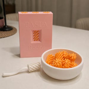Peut inclure: Une boîte rectangulaire rose étiquetée PASTA Rotini avec une fenêtre découpée montrant des pâtes rotini orange. À côté se trouve un bol blanc rempli de rotini orange et un ustensile à manche blanc avec une tête en spirale.
