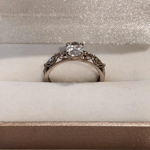 May include: A silver-toned ring featuring a central clear, oval-cut gemstone set in a prong setting. The band displays intricate, openwork metal detailing along the sides, with smaller clear stones embedded within the filigree-style metalwork.