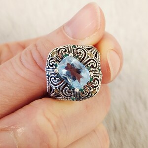 May include: A silver ring featuring a large, rectangular, light blue faceted gemstone at the centre. The band is crafted with intricate, dark-oxidised scrollwork patterns and is accented with small, round, green and yellow gemstones.