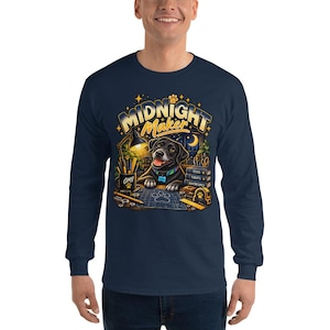 May include: A navy blue long-sleeve t-shirt featuring a graphic of a black dog at a desk with the text "MIDNIGHT MAKER". The illustration includes a lamp, a mug labeled "OMI", books titled "IDEAS", "TREATS", and "ADVENTURES", and tools.