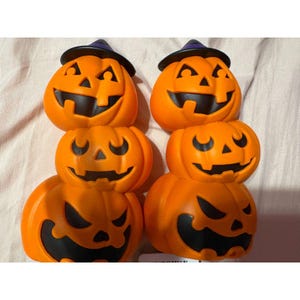 May include: Two vertical stacks of three orange plastic jack-o'-lanterns. Each stack is topped with a small black witch hat featuring a purple band. The pumpkins have carved black facial features including triangular eyes and jagged mouths.