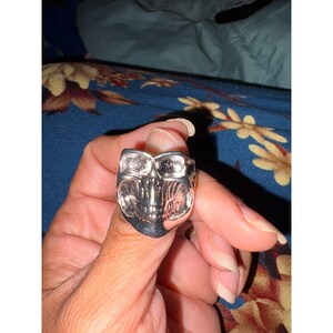 May include: A silver-toned ring featuring a detailed skull design. The skull has prominent eye sockets, a defined nasal cavity, and visible teeth. The metal surface has a polished finish with recessed areas that create depth and contrast.
