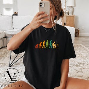 Pu&ograve; includere: Una t-shirt nera a maniche corte con una stampa grafica colorata che raffigura le fasi dell'evoluzione umana, progredendo da un primate a una persona seduta a una scrivania che lavora al computer, disposta in una sequenza orizzontale.