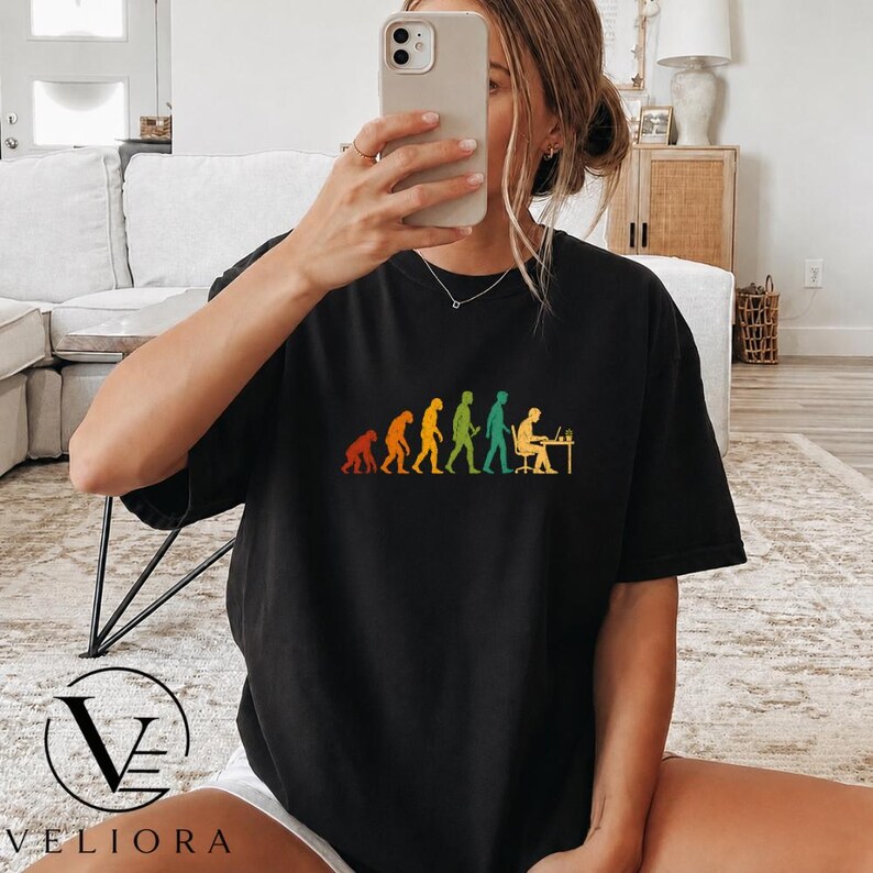 Pu&ograve; includere: Una t-shirt nera a maniche corte con una stampa grafica colorata che raffigura le fasi dell'evoluzione umana, progredendo da un primate a una persona seduta a una scrivania che lavora al computer, disposta in una sequenza orizzontale.