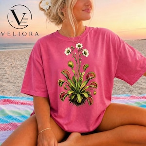 May include: A pink short-sleeved t-shirt featuring a central graphic print of a green Venus flytrap plant with white flowers. The plant illustration displays multiple open traps with red interiors and spiky edges, centred on the chest area.