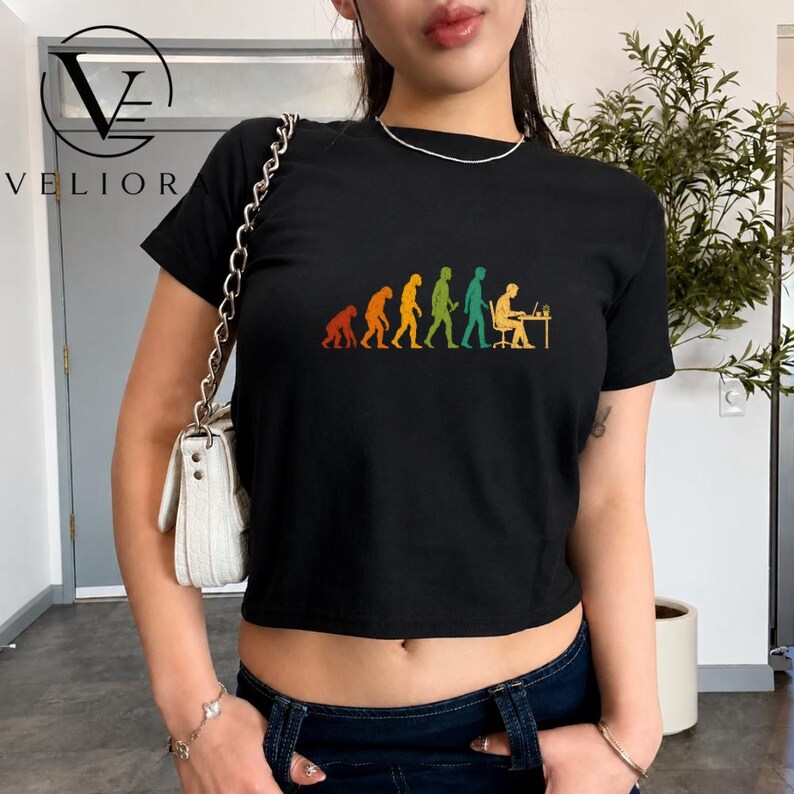 Pu&ograve; includere: Una t-shirt nera a maniche corte con una stampa grafica colorata che raffigura l'evoluzione dell'umanit&agrave;, partendo da un primate fino a una figura seduta a una scrivania con un computer. La maglietta presenta un girocollo e un taglio corto.