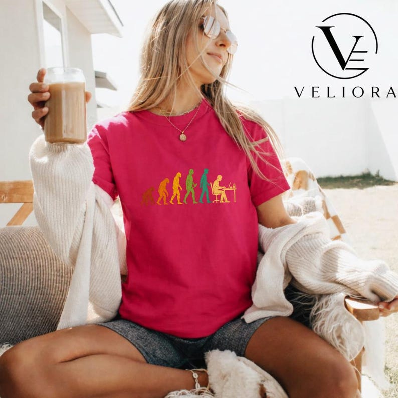 Pu&ograve; includere: Una t-shirt a maniche corte di colore rosa acceso con una stampa grafica dell'evoluzione umana, che progredisce da un primate a una persona seduta a una scrivania con un computer. Il grafico utilizza colori giallo, arancione e verde.