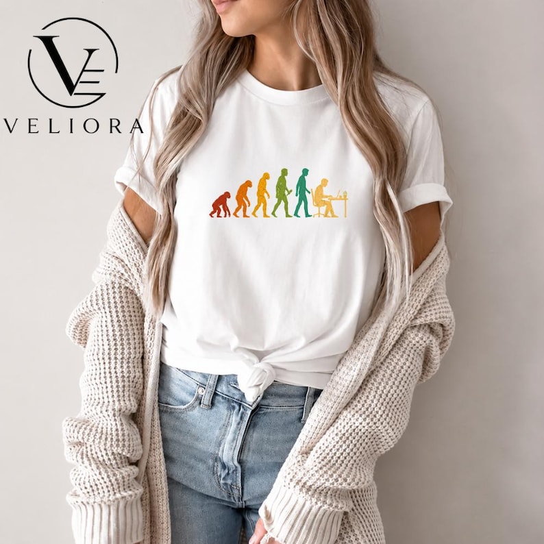 Pu&ograve; includere: Una t-shirt bianca a maniche corte con una grafica colorata dell'evoluzione umana, che progredisce da un primate a una persona seduta a una scrivania con un computer. La maglietta &egrave; indossata sotto un cardigan in maglia beige aperto.