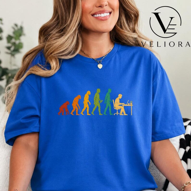 Pu&ograve; includere: Una t-shirt blu reale con una stampa grafica multicolore che mostra l'evoluzione umana, partendo da un primate fino a una persona seduta a una scrivania che lavora su un computer portatile. Il testo VELIORA &egrave; stampato in nero in alto a destra.