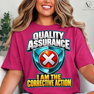 May include: A bright pink t-shirt featuring a graphic with the text "QUALITY ASSURANCE" in white letters above a blue shield icon containing a red circle with a white "X". Below the shield, the text "I AM THE CORRECTIVE ACTION" is printed.