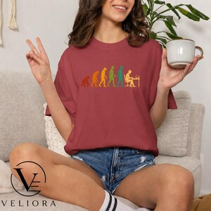 Pu&ograve; includere: Una t-shirt a maniche corte color bordeaux con una stampa grafica multicolore che raffigura l'evoluzione umana, partendo da un primate fino a una persona seduta a una scrivania che lavora al computer, con una tazza in ceramica bianca vicina.