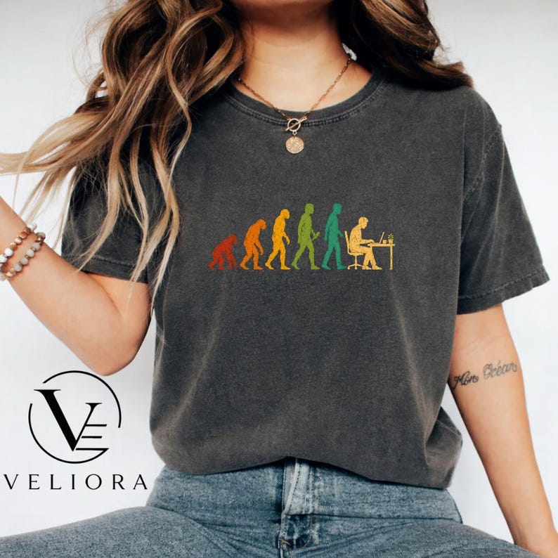 Pu&ograve; includere: Una t-shirt grigio antracite a maniche corte con una stampa grafica dell'evoluzione umana, che progredisce da un primate a una persona seduta a una scrivania con un computer. La grafica utilizza colori arancione, giallo e verde. Girocollo.