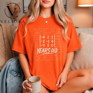 Puede incluir: Una camiseta naranja de manga corta con un estampado gráfico blanco de una matriz matemática y el texto YEARS OLD centrado debajo. El diseño está rodeado por varias fórmulas matemáticas y formas geométricas en color blanco tenue.