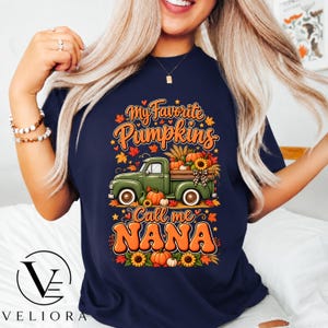May include: A navy blue t-shirt featuring a graphic of a green pickup truck carrying pumpkins and sunflowers. The text reads My Favorite Pumpkins Call me NANA, surrounded by autumn leaves. The design uses orange, yellow, and green colors.
