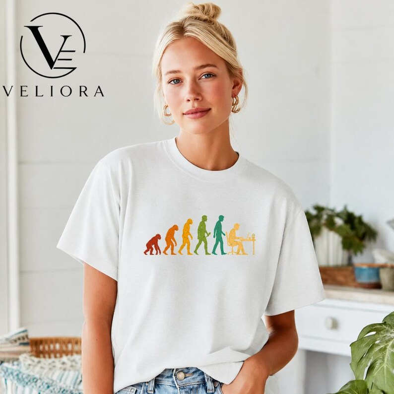 Pu&ograve; includere: Una t-shirt bianca a maniche corte con una stampa grafica colorata dell'evoluzione umana, che inizia con un primate e progredisce fino a un essere umano seduto a una scrivania con un computer. Il design utilizza sagome arancioni, gialle e verdi.