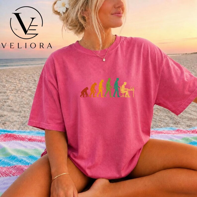 Pu&ograve; includere: Una t-shirt rosa a maniche corte con una stampa grafica multicolore che raffigura l'evoluzione dell'umanit&agrave;, partendo da un primate e progredendo attraverso varie fasi fino a una persona seduta a una scrivania davanti a un computer.