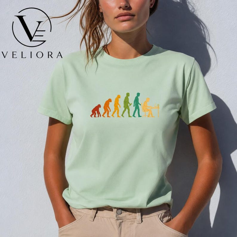 Pu&ograve; includere: Una t-shirt a maniche corte color verde chiaro con una stampa grafica multicolore che raffigura le fasi dell'evoluzione umana, partendo da un primate fino a una figura umana seduta a una scrivania che lavora al computer.