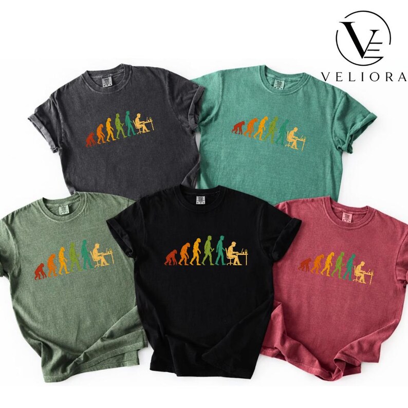 Pu&ograve; includere: Cinque t-shirt a maniche corte con girocollo in grigio antracite, verde, oliva, nero e bordeaux. Ogni maglietta presenta una stampa grafica multicolore che mostra l'evoluzione umana, dai primati a una persona seduta a una scrivania.