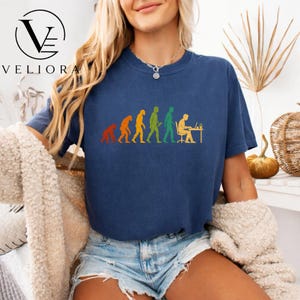 Pu&ograve; includere: Una t-shirt blu navy a maniche corte con una stampa grafica dell'evoluzione umana, che parte da un primate e progredisce fino a una persona seduta a una scrivania con un computer. Il design utilizza i colori arancione, giallo e verde.
