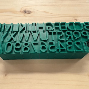 Può includere: Un set di timbri stampati in 3D di colore verde scuro che presentano l'intero alfabeto in lettere maiuscole e i numeri da zero a nove. Ogni carattere è in rilievo su una base a blocco rettangolare, disposti in tre file per una chiara visibilità.