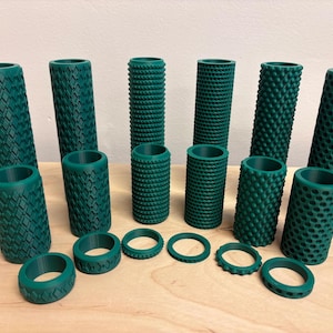 Puede incluir: Una colección de rodillos cilíndricos y anillos a juego de color verde oscuro con diversas texturas geométricas, incluyendo patrones de diamantes, puntos y rejillas, dispuestos en tres filas de alturas variadas.