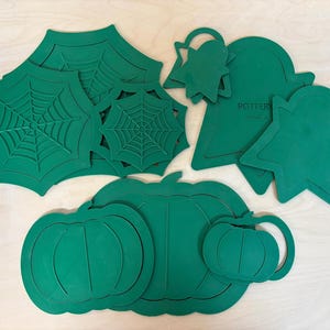 May include: A collection of green silicone mats in various shapes including spider webs, ghosts, and pumpkins. The items feature embossed outlines of their respective designs. One ghost-shaped mat has the word POTTERY printed on its surface.