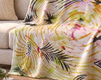 May include: A soft throw blanket featuring a white base with a vibrant watercolor-style print. The design includes abstract yellow and orange circular brushstrokes, pink splatters, and bold black palm leaf patterns throughout.