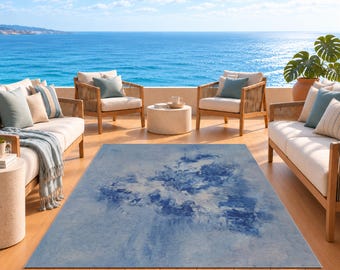 May include: A rectangular area rug featuring a blue and white abstract watercolor-style pattern. The rug is positioned on a wooden deck between two outdoor sofas and two armchairs, all with light-colored cushions and blue accent pillows.