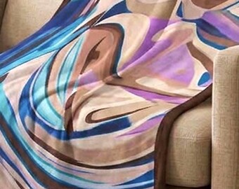 May include: A rectangular throw blanket featuring an abstract pattern of swirling blue, purple, tan, and white colors. The blanket has a solid dark brown border along all four edges and is draped over the seat and back of a beige sofa.