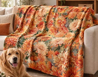 May include: A soft throw blanket featuring a dense, repeating pattern of vibrant orange, yellow, and cream-colored dahlia flowers set against a deep green foliage background, draped over the back and seat of a light-colored sofa.