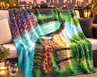 May include: A soft throw blanket featuring a vibrant, painterly landscape print of a cascading waterfall surrounded by lush green foliage and clusters of bright purple, pink, and yellow flowers, draped over a neutral-colored outdoor sofa.