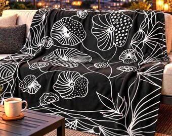 May include: A black throw blanket featuring a white line-art pattern of stylized lotus flowers and lily pads. The design includes circular shapes with dotted textures and radiating lines, creating a continuous botanical motif across the fabric.